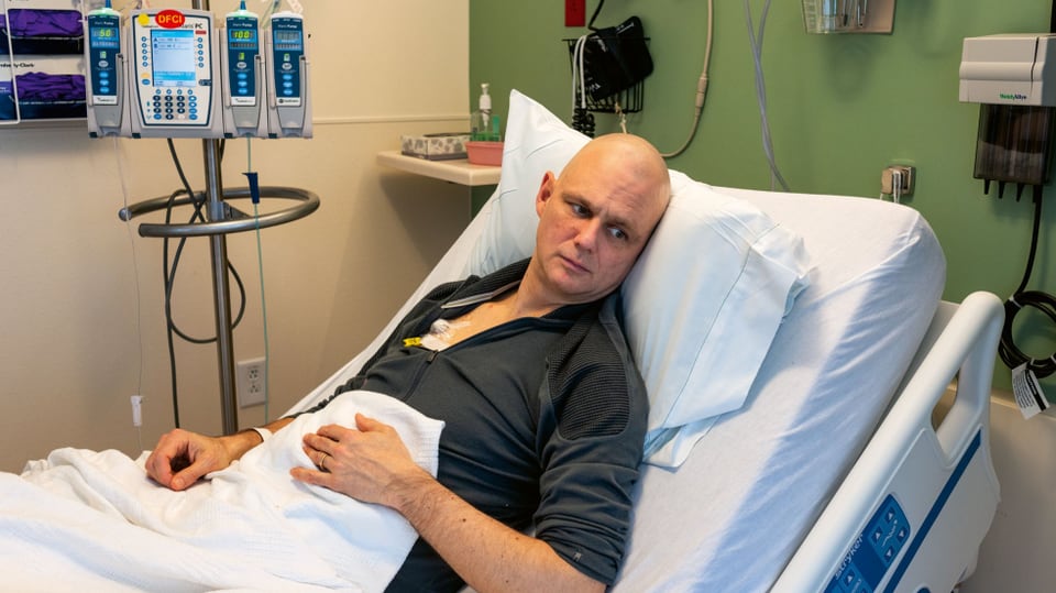 Ein Mann liegt mit dunklem Pullover in einem Spitalbett.