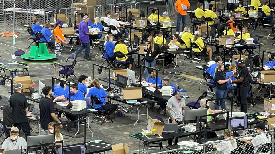 Tische mit Menschen in verschiedenfarbigen T-Shirts, die Wahlzettel zählen und untersuchen