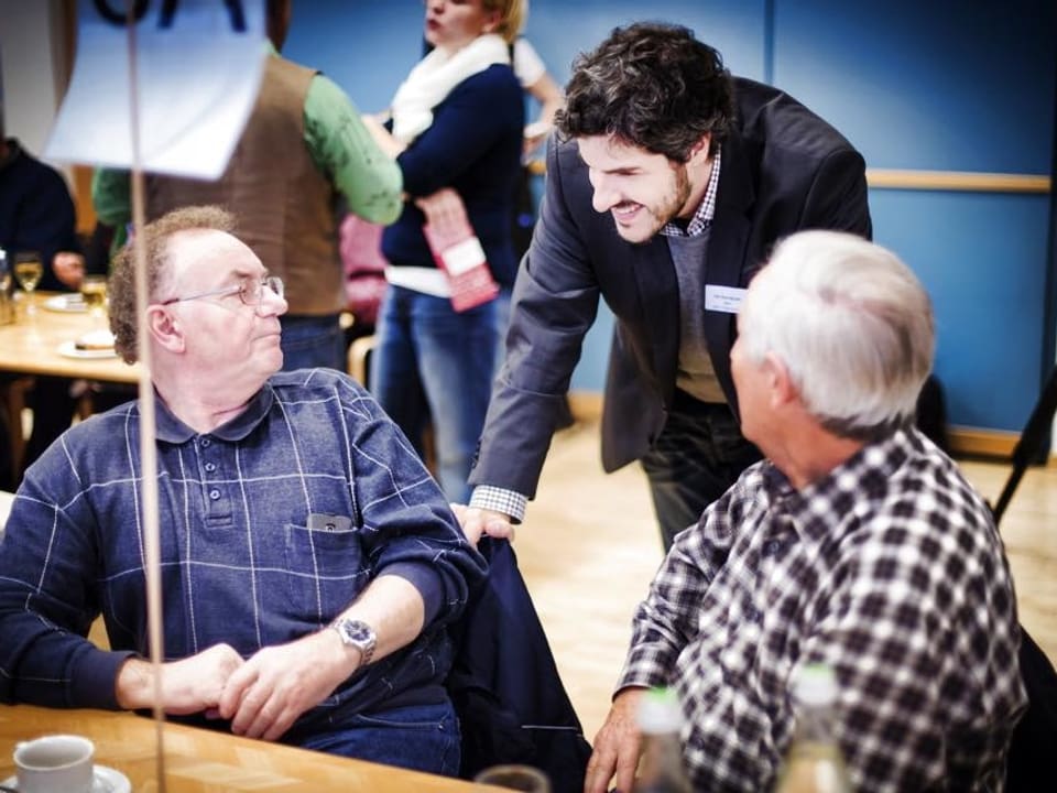 Zwei ältere Männer sitzen an einem Tisch, ein junger Mann beugt sich zu ihnen und spricht mit ihnen.