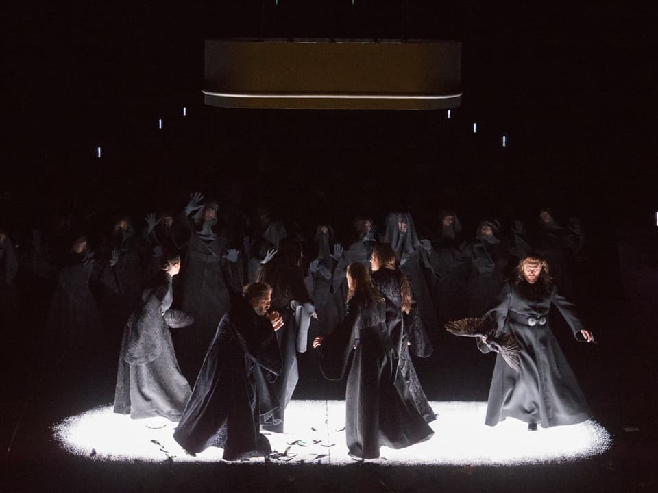 Männer und Frauen in grauen Kostümen tanzen über eine Bühne.