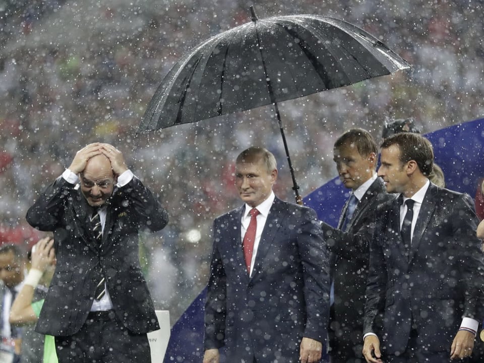Macron und Infantino werden direkt vom Regen erfasst, während Putins Angestellter ihm den Schirm hinhält.
