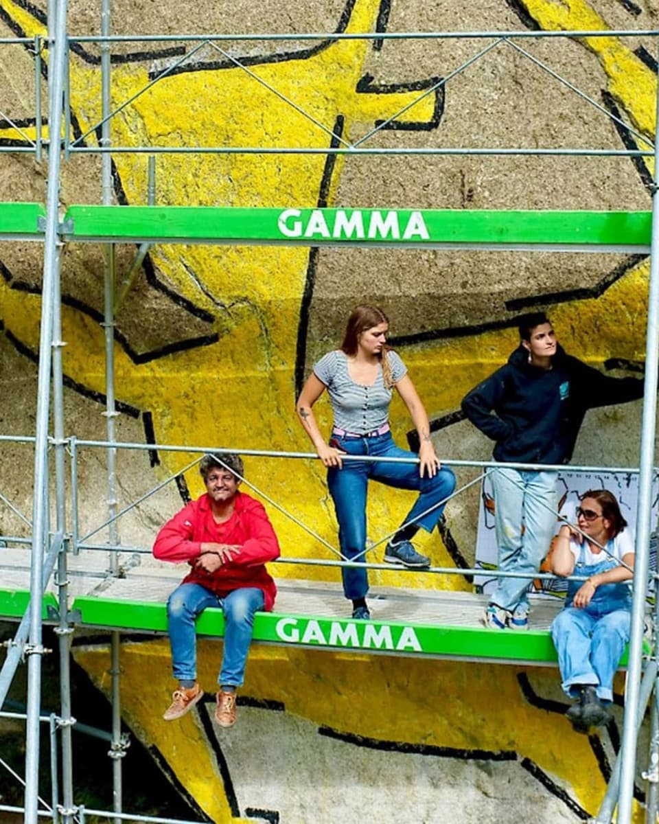 Vier Menschen stehen und sitzen auf einem Baugerüst an einer Felswand, auf der ein grosser gelber Teufel aufgemalt ist.