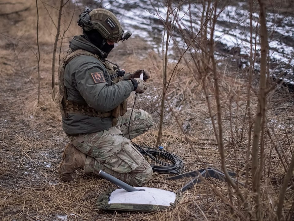 Ein Soldat sitzt im Tarnanzug am Boden und hantiert mit einem Kabel.