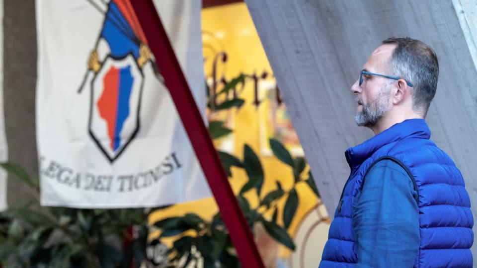 Norman Gobbi steht seitlich. Im Hintergrund hängt die Tessiner Kantons-Flagge.