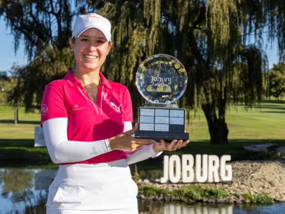 Golferin hält einen Pokal vor einem Teich mit der Aufschrift 'Joburg'.