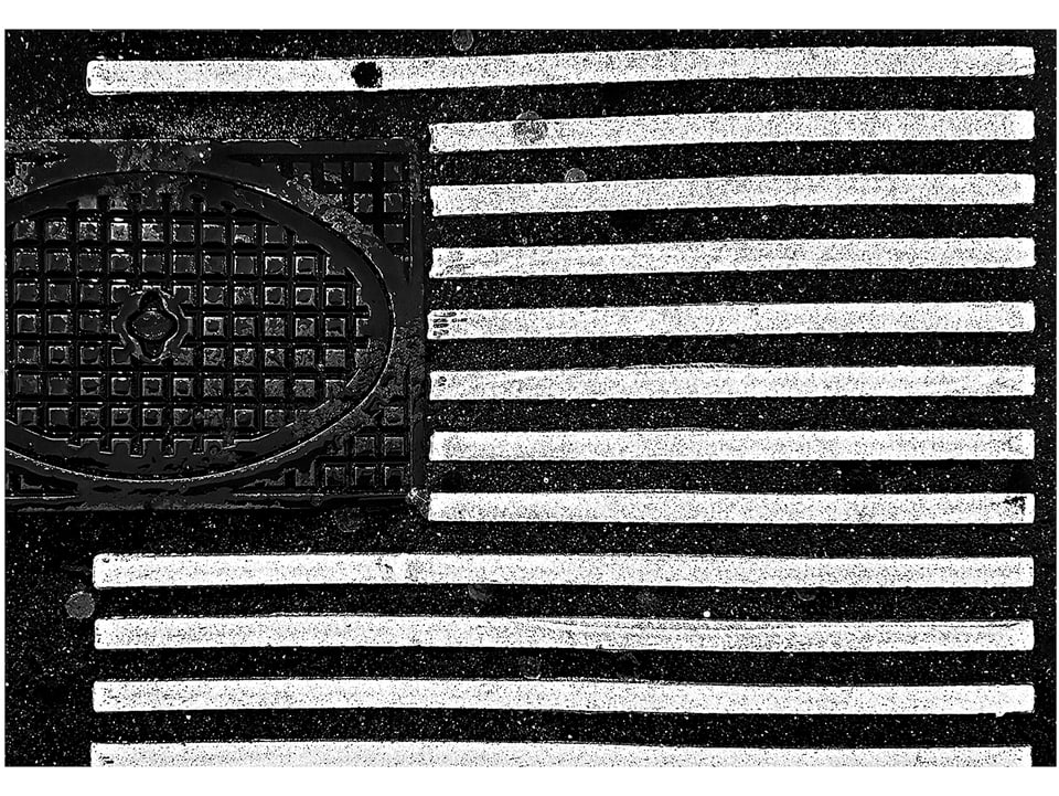 Ein Gulli und weisse Bodenstreifen für Blinde. Die Bildkomposition lässt den Boden wie die US-Flagge erscheinen.
