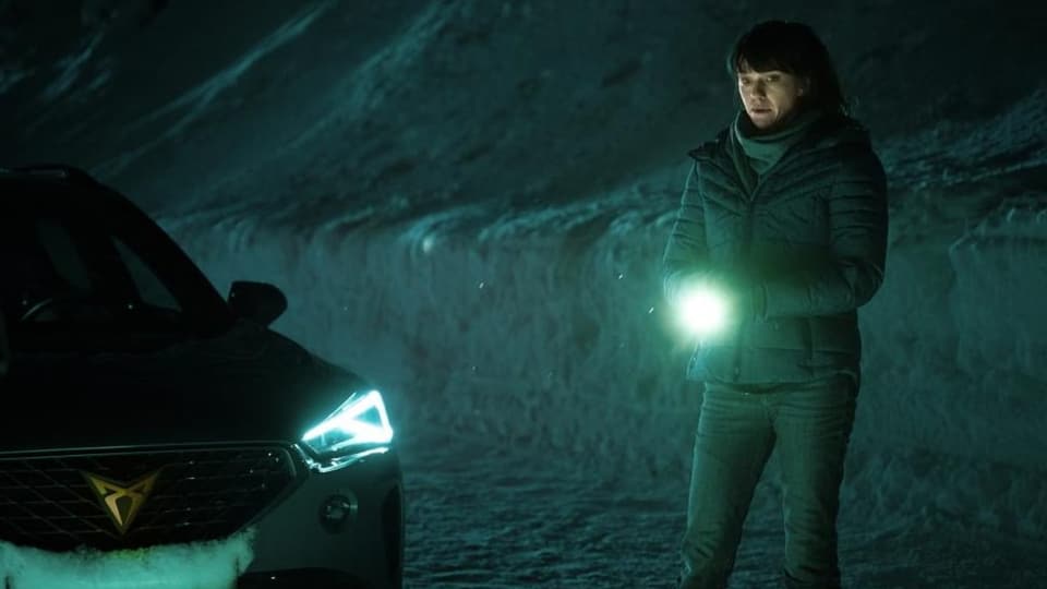 Foto, Frau steht in der Nacht draussen im Schnee mit Taschenlampe, links ein Auto mit Vorderlicht.