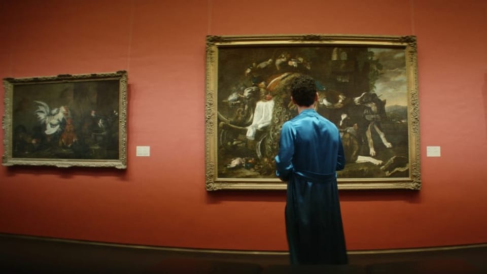Mann in blauem Kimono steht vor roter Wand mit hängenden Kunstwerken.