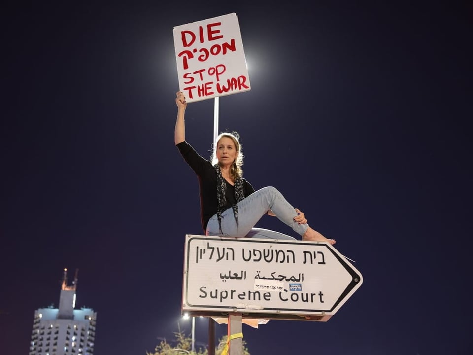 Frau auf Strassenschild. Sie hält Plakat hoch. Darauf steht: Stoppt den Krieg.