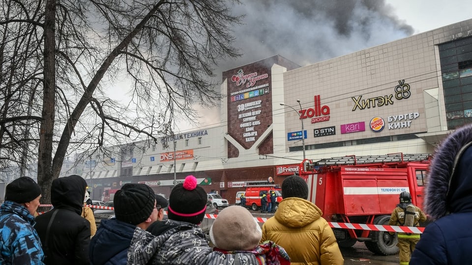Menschen stehen hinter einer Absperrung vor dem brennenden Einkaufscenter.