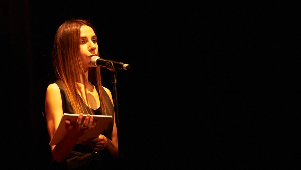 Frau auf einer Bühne vor einem Mikrofon. Sie hält einen Stapel Blätter in der Hand.