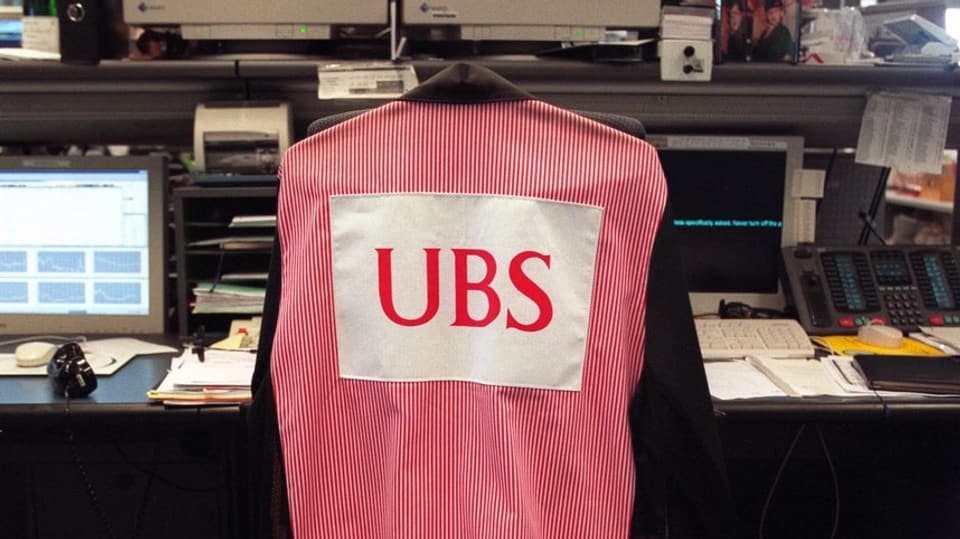 Über einem Stuhl häng eine Jacke mit der Aufschrift UBS. Der Stuhl gehört zu einem Arbeitsplatz in einer Bank.
