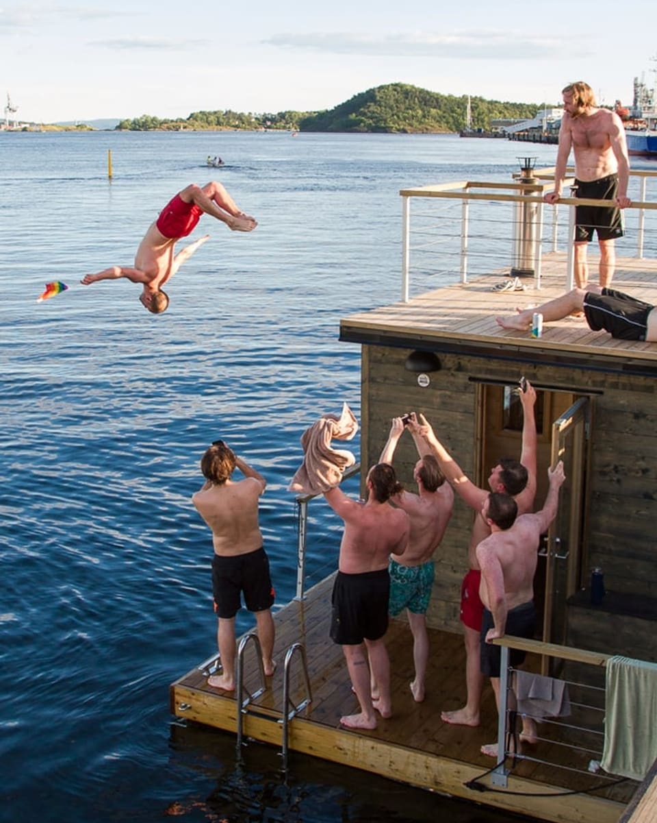 Ein Mann springt rückwärts von einem Hausboot ins Wasser. Seine Freunde jubeln ihm zu.