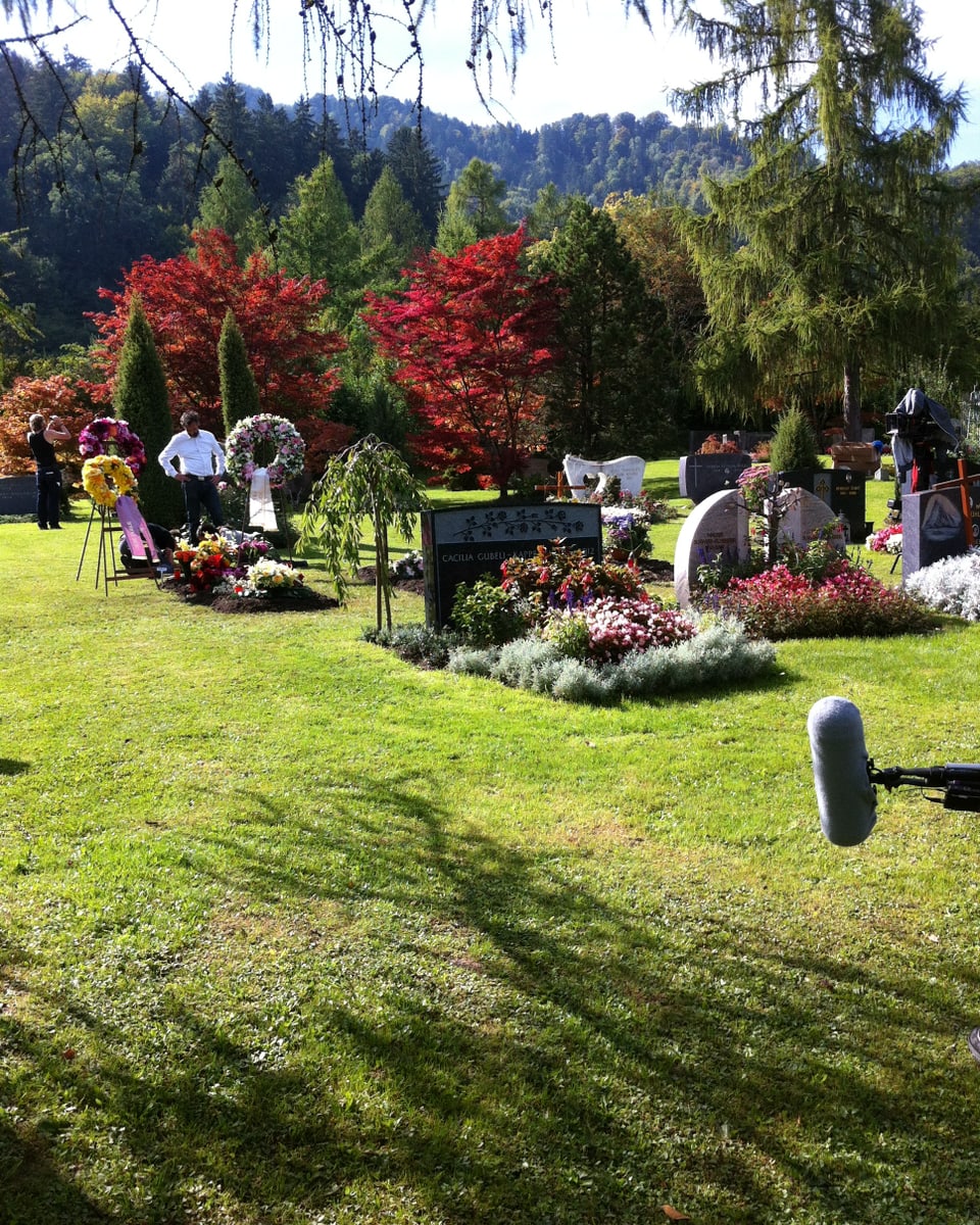 Ein Friedhof, der wie bei einer Beerdigung geschmückt ist. Die Sonne scheint in vollen Zügen. Am rechten Bildrand ist ein Mikrofon zu sehen.