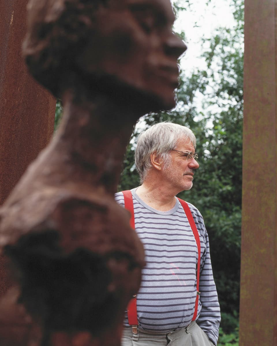 Der Künstler Schang Hutter gestreiftem Pulli und Hosenträgern steht hinter einer rostigen Skulptur.
