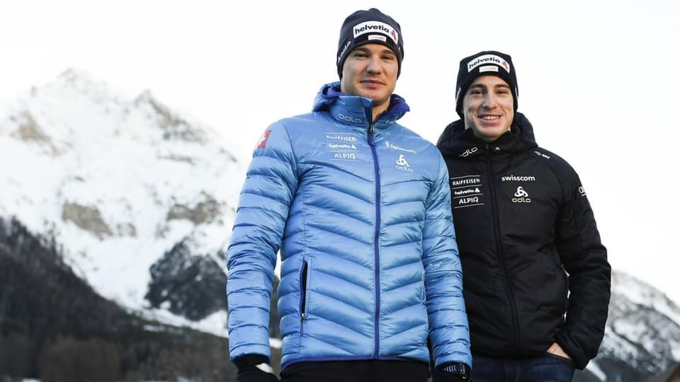 Zwei Männer in Wintersportkleidung lächelnd vor einem verschneiten Berg.