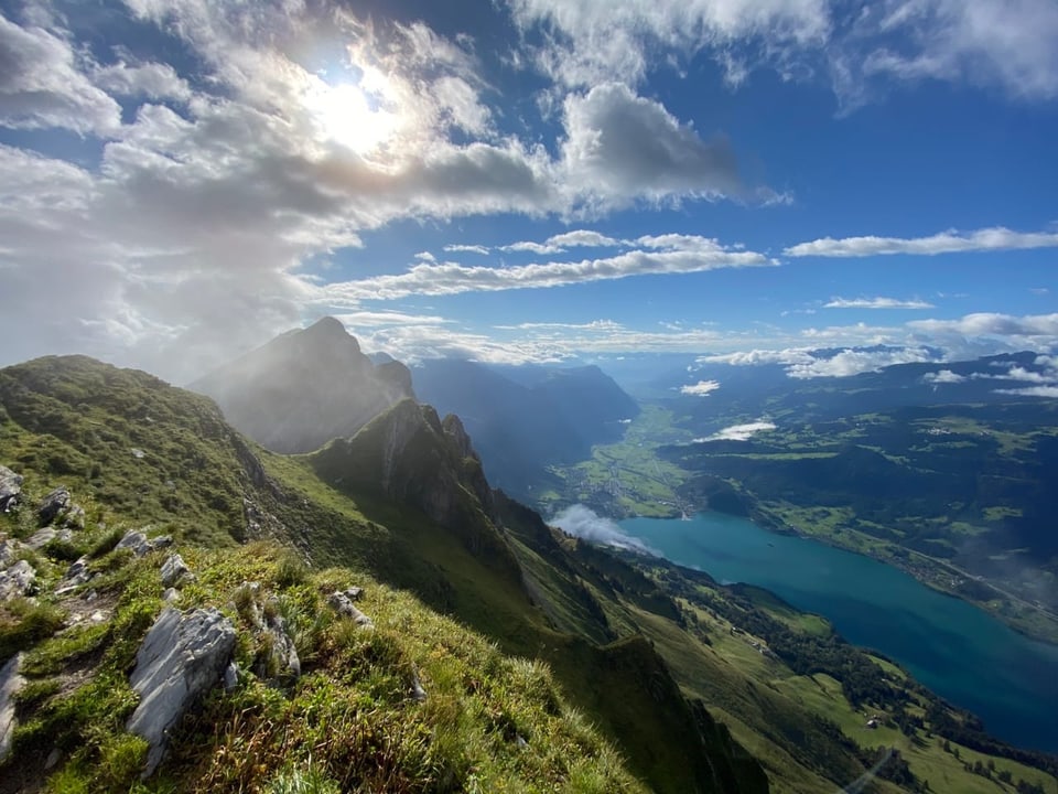 Blick von einer Bergkette mit grünen Wiesen und Felsen auf einen See herunter. Der Himmel ist leicht bewölkt. 