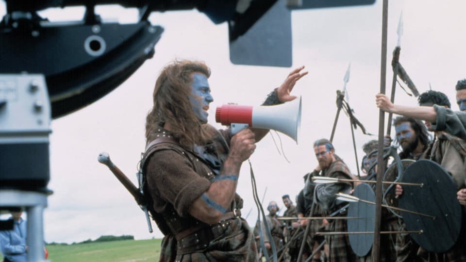 Ein Mann mit langen Haaren, Schottenrock und einem Megafon in der Hand steht vor einer Horde verkleideter Männer und gibt Anweisungen.