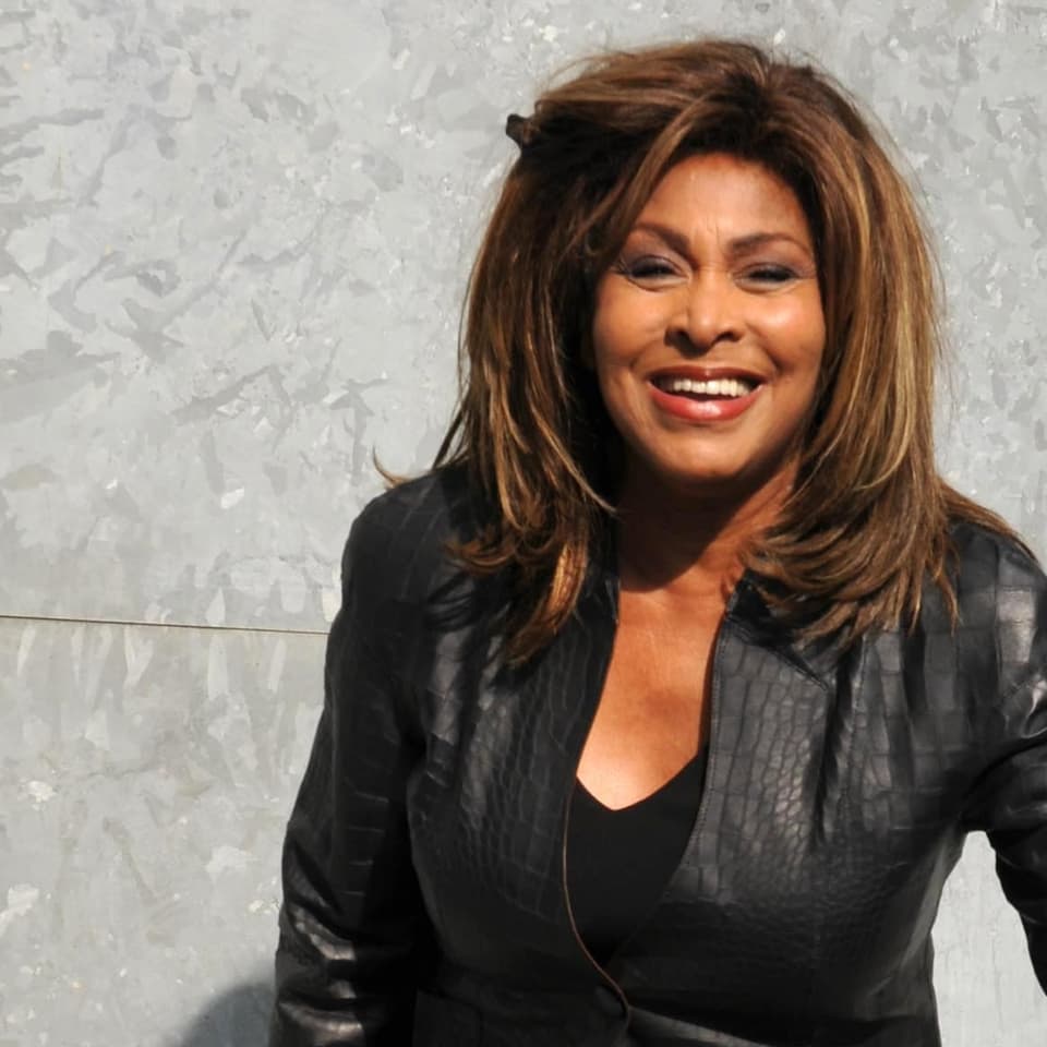 Tina Turner posiert strahlend ganz in schwarz gekleidet mit Lederjacke.