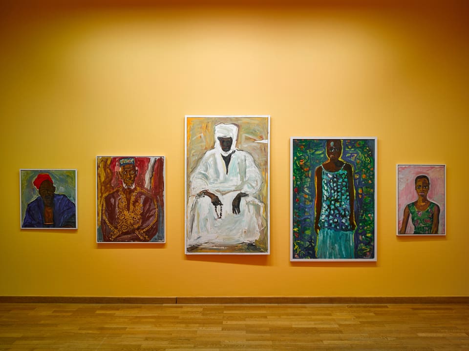 An einer gelb gestrichenen Wand hängen fünf unterschiedlich grosse Bilder. Alles Porträts von afrikanischen Frauen und Männern.