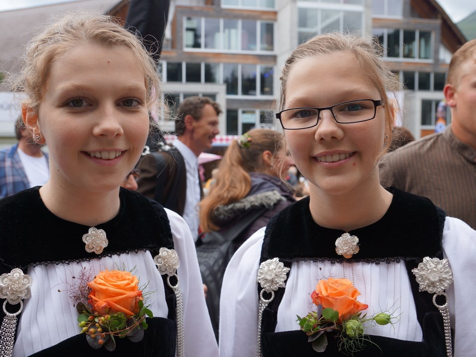 Zwei junge Mädchen mit Trachten und hübschem, orangefarbenem Rosengesteck am Decolletée.