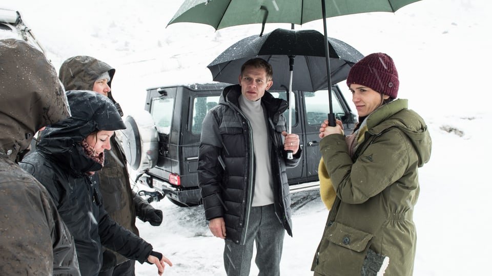 Die beiden Schauspieler stehen mit Schirmen in einer verschneiten Landschaft.