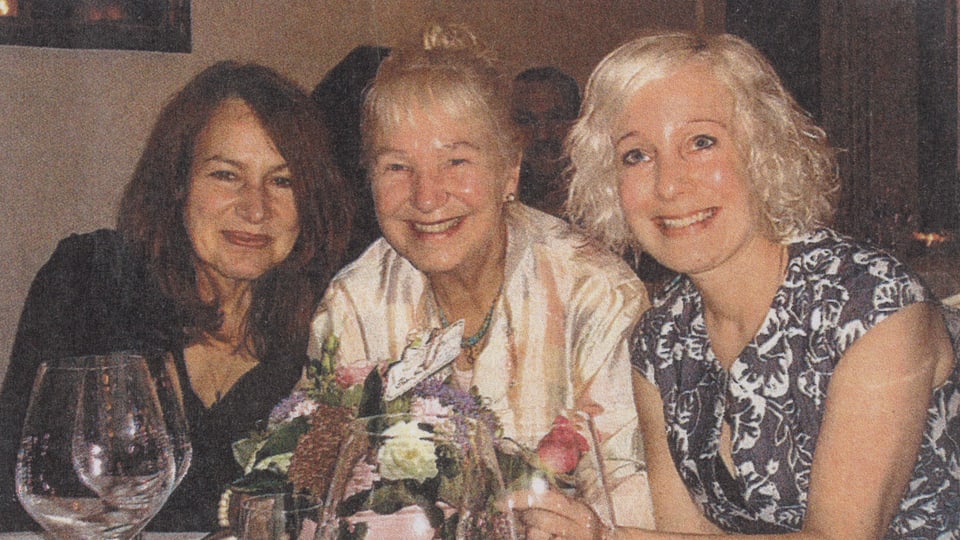 Drei Frauen sitzen an einem Tisch und lächeln in die Kamera