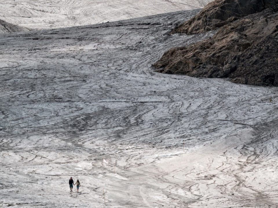 Weitwinkelaufnahme eines Gletschers, der sich um einen Berg schmiegt. In der Mitte des Bildes laufen zwei Menschen.
