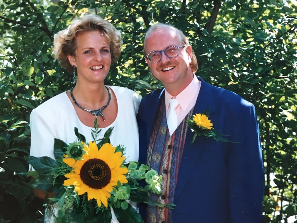 Ein lächelndes Brautpaar, sie in weiss, er in blauem Anzug mit farbiger Weste. Sie trägt eine Sonnenblume in der Hand, er eine gelbe Blume im Knopfloch.