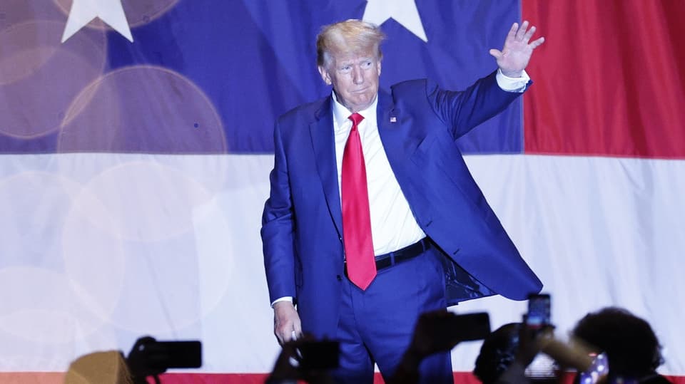 Donald Trump steht im Anzug und mit einer roten Krawatte auf einer Bühne und winkt der Menge zu.