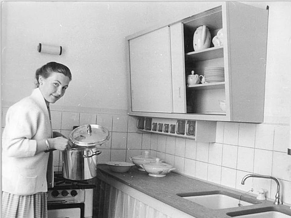 Ein Frau hebt den Deckel eines Kochtopfs und schaut in die Kamera.