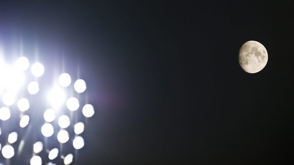 Flutlichter eines Fussballstadions leuchten in der Nacht, am Himmel der Mond.