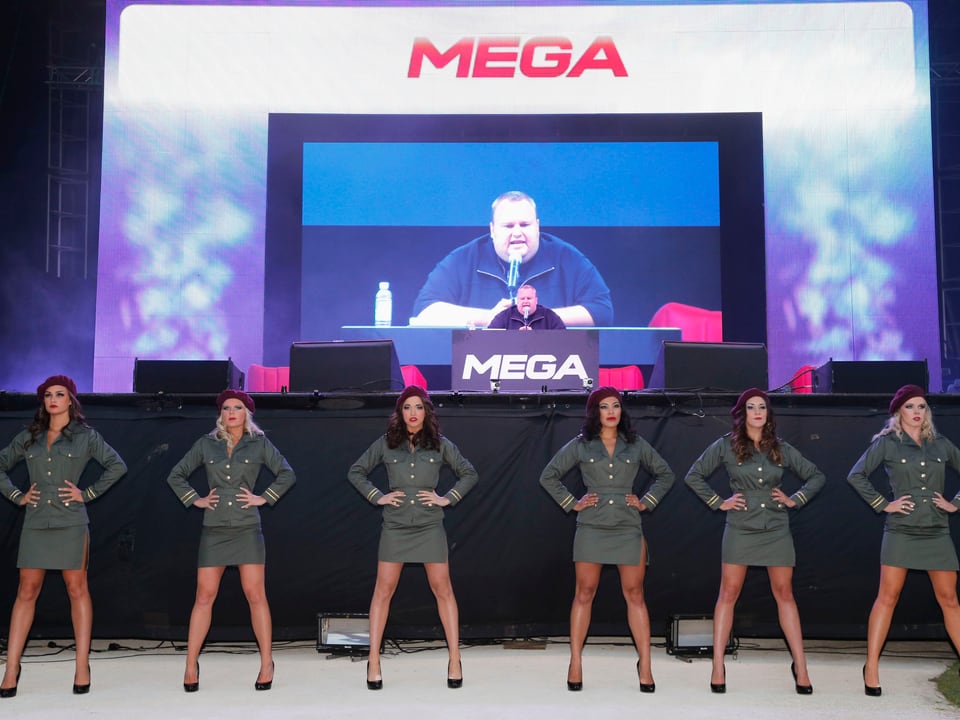 Sechs Tänzerinnen in Militäruniform und kurzem Rock stehen vor einer Videoleinwand, auf der Kim Dotcom zu sehen ist, wie er in ein Mikrophon spricht.
