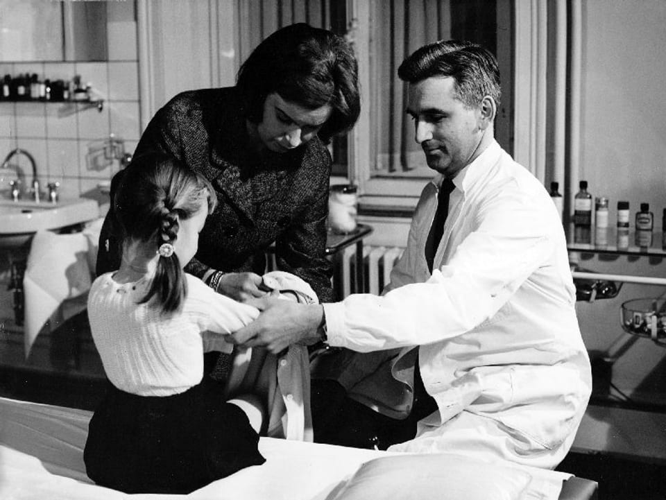 Ein Arzt im Kittel sitzt vor einer Liege und gibt dem Mädchen, welches auf der Liege sitzt eine Spritze. Zwischen ihnen steht eine Frau, die mit einem Tuch bereitsteht.