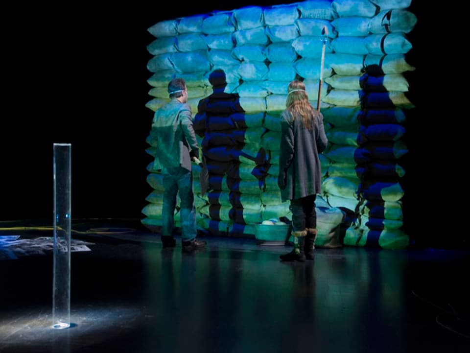 Szene aus einem Theaterstück. Vier Leute stehen vor einer Wand aus Sandsäcken.