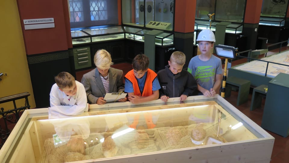 Fünf Jungs schauen in eine Glasvitrine, in welcher sich ein 2000 Jahre alter Grabstein befindet. Im Hintergrund ist das Museum zu sehen.