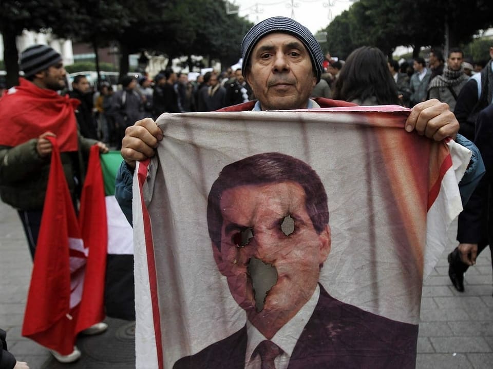 Protestierender mit einer Fahne mit dem Gesicht von Zine el-Abidine Ben Ali, Augen und Mund sind ausgestochen.