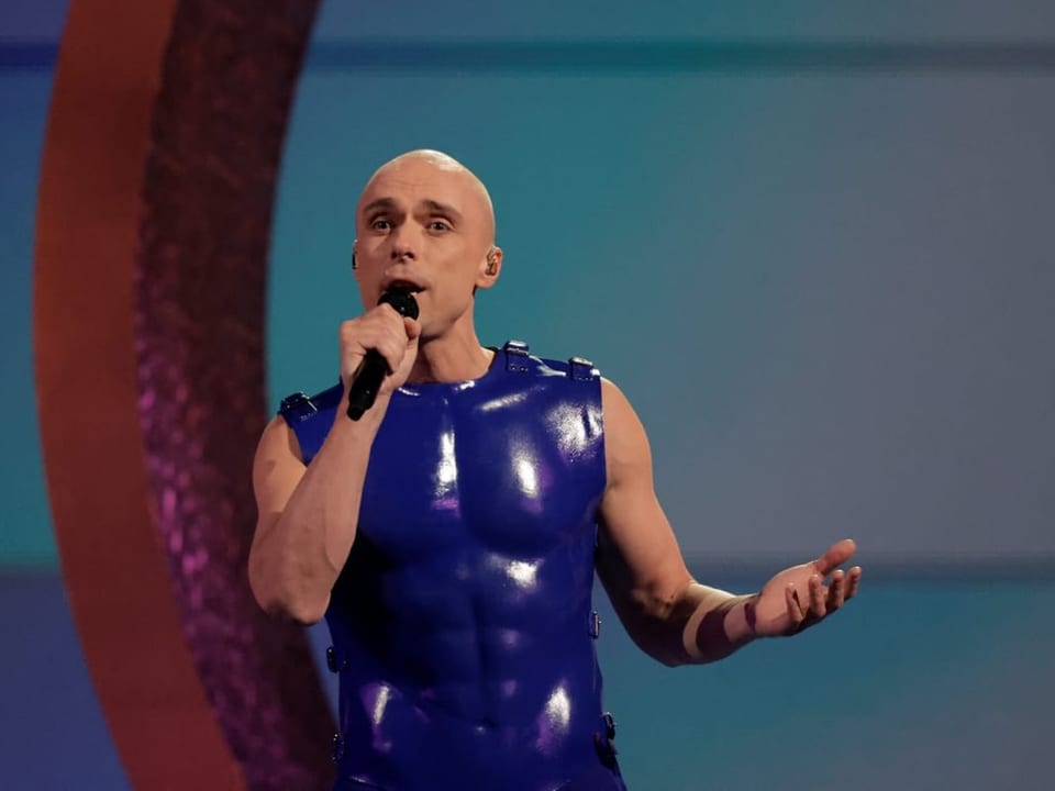 Glatzköpfiger Mann in blauem Latex-Outfit singt in ein Mikrofon auf einer Bühne.