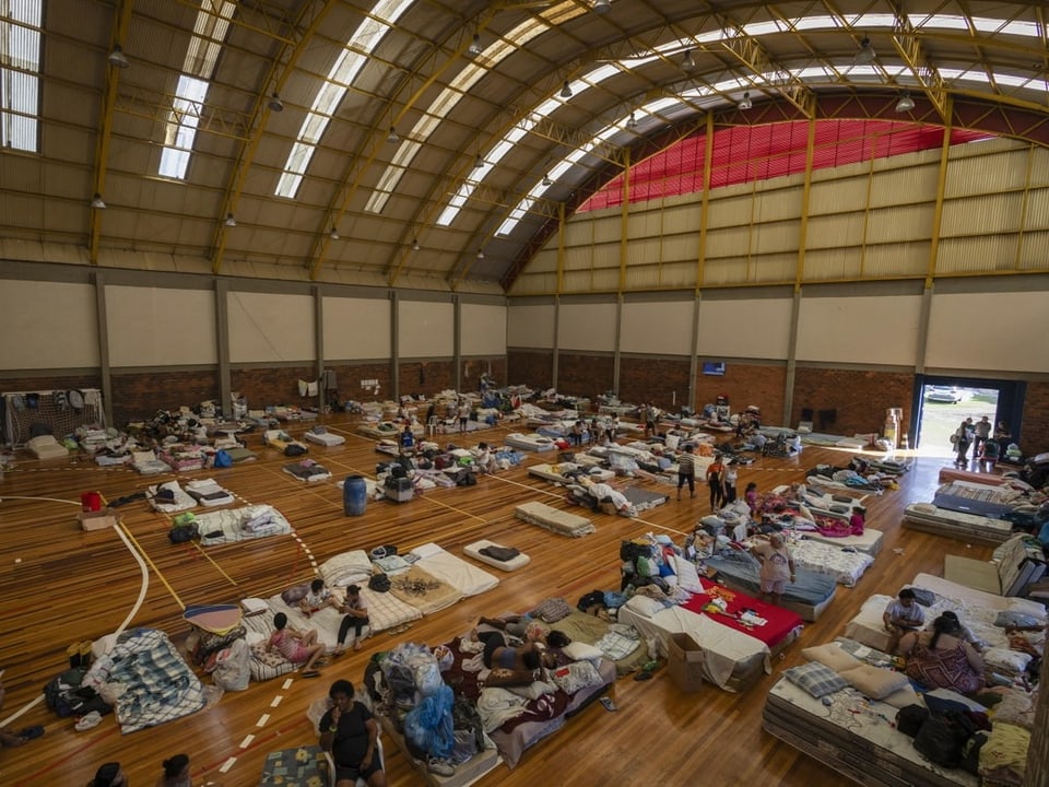 Eine Notunterkunft in einer Sporthalle mit einem gekrümmten Dach. Auf dem Boden liegen Menschen auf flachen Mattratzen.