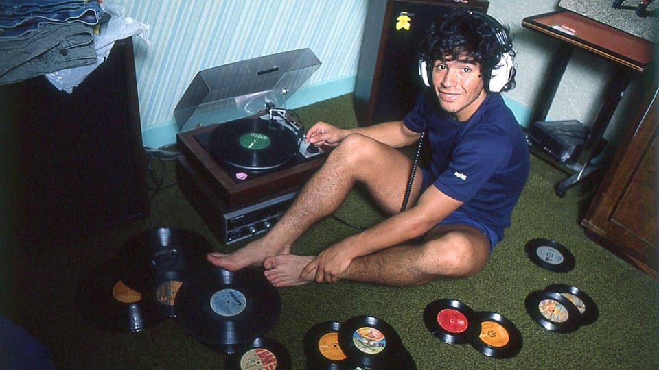 Ein junger Maradona sitzt auf dem Fussboden neben einem Plattenspieler. Um ihn herum sind viele Vinyl-Platten, er hat Kopfhörer auf.