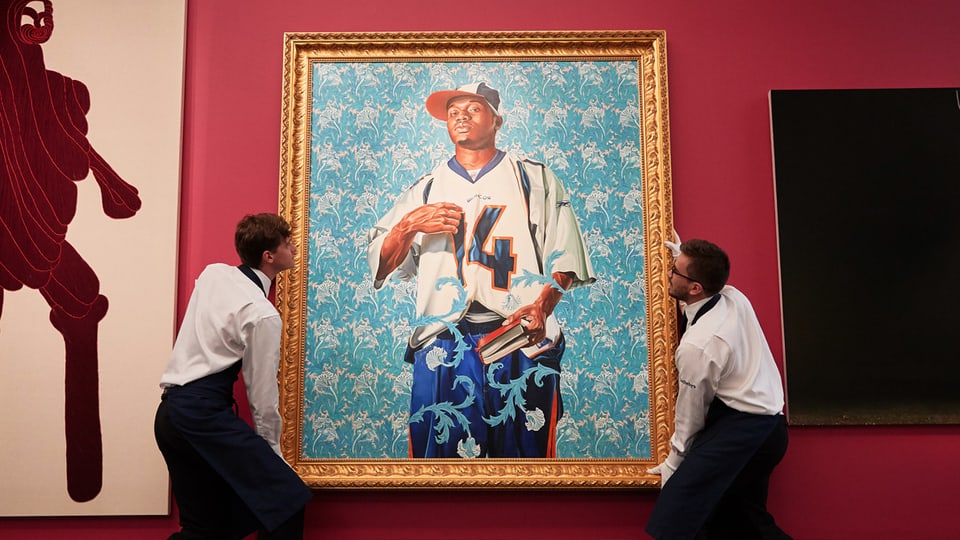 Ein gerahmtes Bild zeigt einen jungen schwarzen Mann in Hip-Hop-Kleidung und mit einem Buch in der Hand. Zwei Mitarbeiter eines Museums hängen es auf.