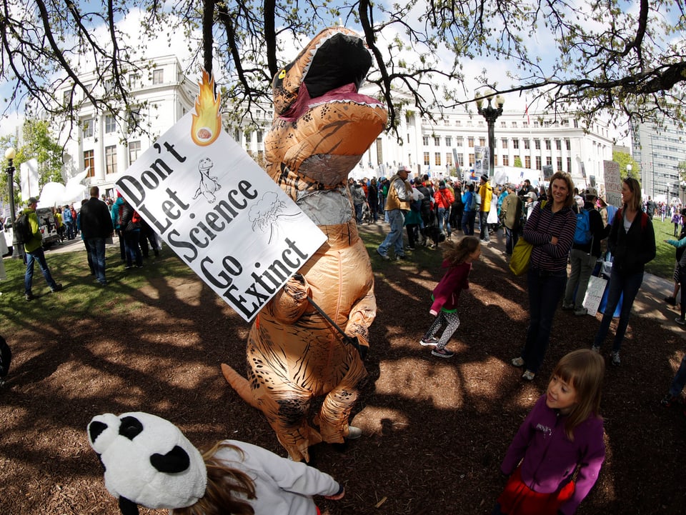 Ein als Dinosaurier verkleidete Person hält ein Plakat, auf dem steht: «Don't Let Science Go Extinct».