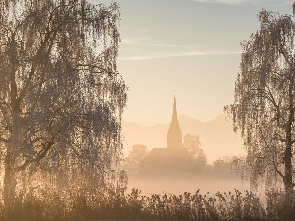 Die Kirche in Kirchdorf ist wie eine Gmälde in sanftes Licht getaucht. Die Morgensonne beleuchtet zudem Bäume und Grässer. Flacher Nebel liegt über den Wiesen.