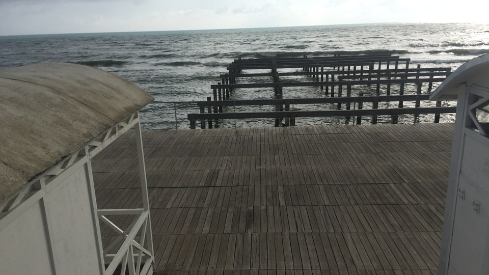 Im Vordergrund die Strandhäuschen auf einer mit Holz bedeckten Plattform, dahinter aus dem Meer ragende Holzpfähle.