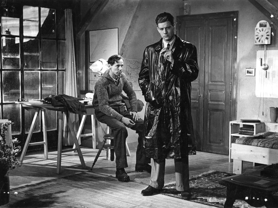 Ein Mann im Ledermantel steht in der Mitte einer Attikwohnung und raucht, hinter ihm sitzt ein zweiter Mann auf einem Holzstuhl.