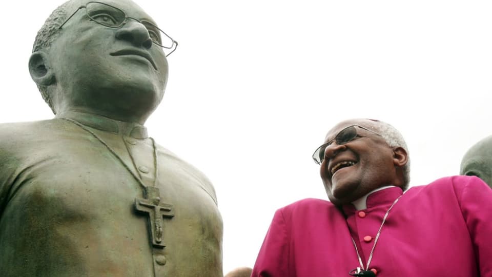 Links steinerne Figur eines Mannes, rechts dunkelhäutiger Bischof in pinkem Gewand, lacht.