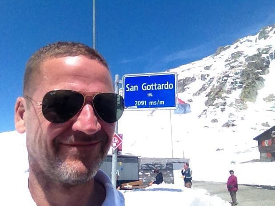 Mann lacht in Kamera, im Hintergrund Schnee und ein Schild "San Gottardo, 2016 müM".