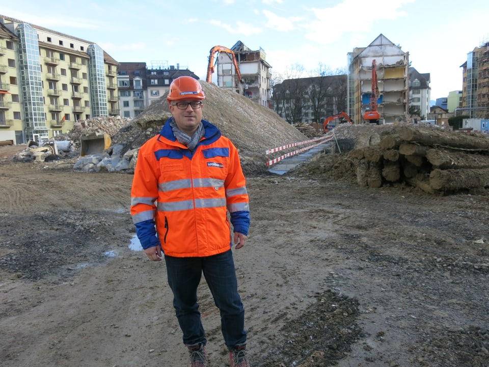 Ein Mann in oranger Leuchtweste und mit Helm auf einer Baustelle.