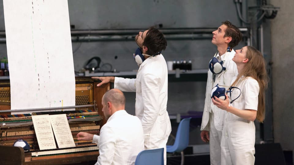 Vier Menschen in weissen Anzügen: Drei stehen und blicken in die Höhe, einer sitzt am Klavier.