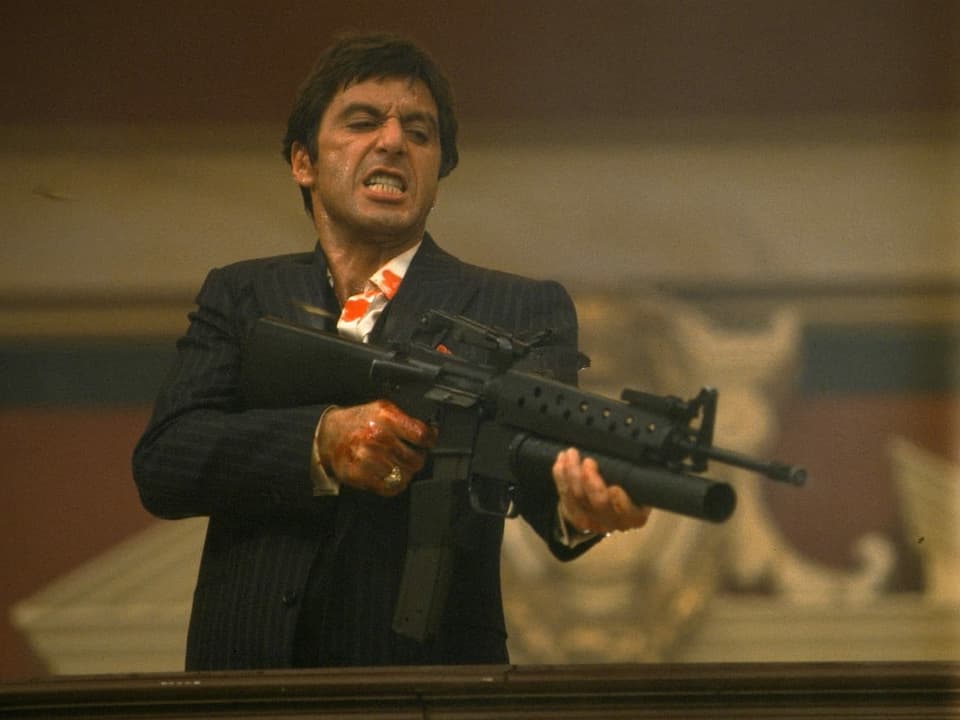 Al Pacino als Tony Montana im Film Scarface. Er trägt einen schwarzen Nadelstreifenanzug. In der Hand hält er ein Maschinengewehr. Er scheint gerade auf jemanden zu schiessen.